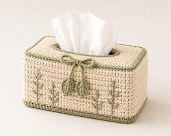 May include: A rectangular crochet tissue box cover in a light beige yarn with a sage green border. The front features embroidered sage green sprigs and a decorative crochet bow with two dangling leaf-shaped accents. A white tissue is visible.