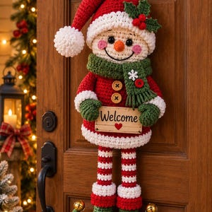 Könnte beinhalten: Ein gehäkelter Schneemann als Wandbehang mit roter Mütze und weißem Bommel, grünem Schal mit Schneeflocke und einem Holzschild mit der Aufschrift Welcome und einem roten Herz. Der Schneemann hat gestreifte Beine und grüne Schuhe mit Goldglocken.