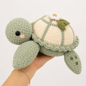 May include: A crocheted turtle plush toy featuring a sage green head and flippers, a cream-colored shell with sage green borders, a small white flower with a yellow center, and a single sage green leaf attached to the top of the shell.