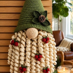May include: A crochet gnome wall hanging featuring a tall green pointed hat adorned with a small pinecone and dark green leaves. The gnome has a round beige nose and a long, curly cream-colored beard decorated with several clusters of red berries.
