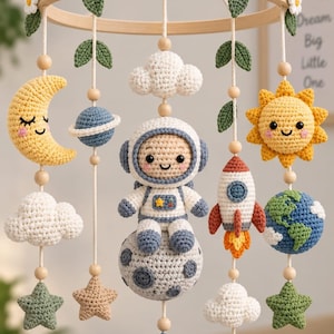 May include: A crochet baby mobile featuring a wooden ring decorated with white flowers and green leaves. Hanging elements include a yellow crescent moon, a blue planet with rings, a white cloud, an astronaut on a moon, a rocket, a sun, and stars.