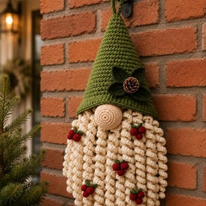 May include: A crocheted gnome wall hanging featuring a tall olive green pointed hat adorned with dark green leaves and a small pinecone. The gnome has a round beige nose and a long, curly cream-colored beard decorated with small red berry clusters.