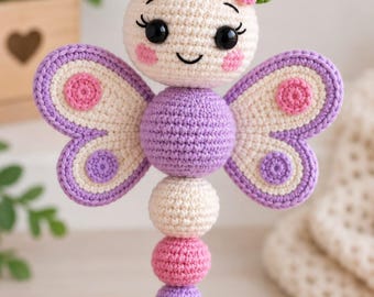 May include: A crochet butterfly rattle featuring a cream-colored head with black eyes, pink cheeks, and a small flower. The body consists of stacked cream and pink spheres, with purple and cream wings decorated with pink and purple circular accents.