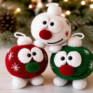 May include: Three round, crocheted ornaments featuring cartoon-like faces with large white eyes, black pupils, and round noses. One ornament is white with red and green accents, one is red with white snowflakes, and one is green with white snowflakes.