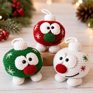 Pode incluir: Três ornamentos redondos de crochê com rostos estilo desenho animado, olhos brancos grandes, pupilas pretas e narizes pequenos. Os ornamentos são vermelhos, verdes e brancos, decorados com padrões de estrelas bordadas e pés brancos.