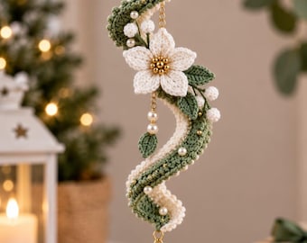 May include: A hanging crochet ornament featuring a wavy, two-toned green and cream yarn structure adorned with small white beads and a central white flower with a gold beaded center. A clear, teardrop-shaped crystal pendant hangs from the bottom.