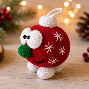 May include: A round, red crocheted ornament featuring a white top loop, two large white eyes with black pupils, a small green nose, and a black stitched mouth. The red body is decorated with white embroidered star-shaped accents and white feet.
