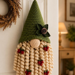 May include: A hanging crochet gnome decoration featuring a tall, pointed olive green hat adorned with a dark green leaf accent and a small pinecone. The gnome has a round beige nose and a long, curly cream-colored beard decorated with small red berries.