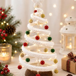 Pode incluir: Uma árvore de Natal em espiral feita de fio de crochê branco, com uma estrela no topo e uma base de crochê marrom. A árvore é decorada com pequenos ornamentos esféricos pendurados em vermelho, verde e dourado, iluminados por luzes.