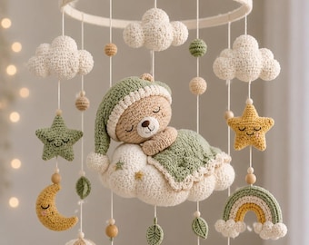 May include: A crochet baby mobile featuring a central sleeping bear wearing a green cap and blanket on a white cloud. Hanging elements include white clouds, yellow stars, a yellow crescent moon, a rainbow, and a hot air balloon in green and beige.