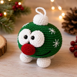 May include: A round, green crocheted ornament featuring two large white eyes with black pupils, a prominent red nose, and a white cap with a hanging loop. The green body is decorated with white stitched snowflake patterns and a black embroidered smile.