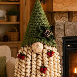 May include: A crochet gnome wall hanging featuring a tall, pointed olive green hat adorned with dark green leaves and a brown pinecone. The gnome has a round beige nose and a long, cream-colored curly beard decorated with small red berries.