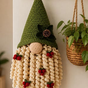 May include: A crocheted gnome wall hanging featuring a tall olive green pointed hat adorned with dark green leaves and a small pinecone. The gnome has a round beige nose and a long, curly cream-colored beard decorated with several clusters of red berries.