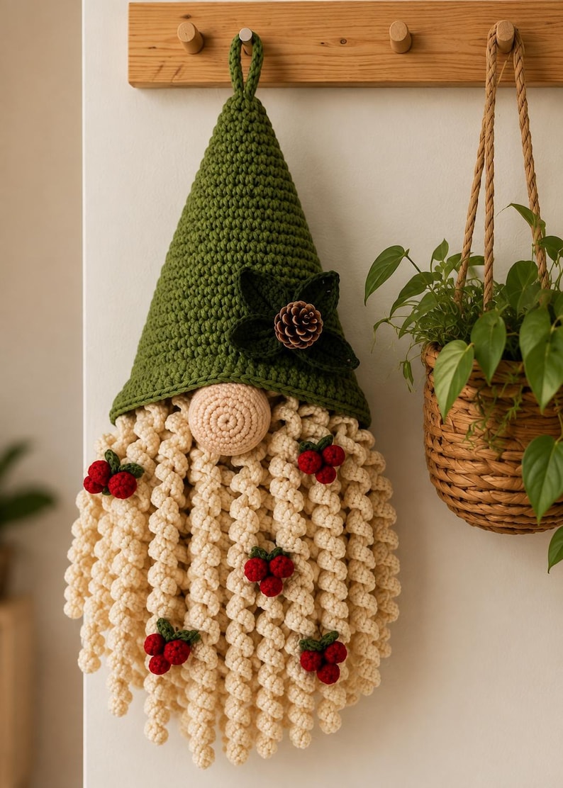 May include: A crocheted gnome wall hanging featuring a tall olive green pointed hat adorned with dark green leaves and a small pinecone. The gnome has a round beige nose and a long, curly cream-colored beard decorated with several clusters of red berries.