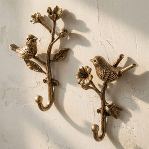 May include: Two antique brass-toned wall hooks, each featuring a sculpted bird perched on a branch with leaves and flowers. The hooks have a curved bottom end and are mounted with visible screws, showcasing a textured, metallic finish throughout.