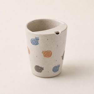 May include: A speckled beige ceramic cup featuring a pattern of small blue, orange, and black cat illustrations. The cup has a unique rim design with a built-in spout or notch on one side, and the exterior surface is textured with tiny dark flecks.