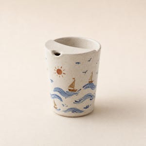 May include: A speckled off-white ceramic cup featuring a hand-painted nautical scene with blue waves, brown sailboats, a small orange sun, and black birds. The cup has a unique rim design with a small integrated spout for pouring liquids.