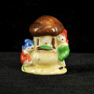 May include: A small ceramic figurine featuring a central cream-colored structure topped with a brown mushroom cap. Two figures are positioned on either side, one wearing a blue hat and red clothing, the other in a red hat and green clothing.