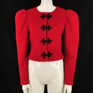 May include: A bright red quilted jacket featuring long sleeves with puffed shoulders and a round neckline. The front closure consists of four black decorative frog fasteners arranged vertically down the center of the garment.