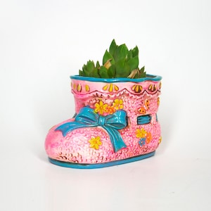 May include: A pink ceramic planter shaped like a baby shoe with a blue bow and floral details. The planter is filled with a green succulent plant.