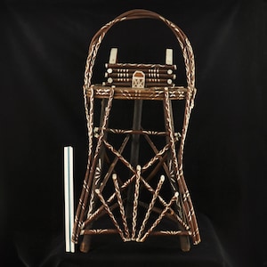 May include: A brown and white decorative basket with a handle. The basket has a tall, openwork design with a geometric pattern. The basket is made of woven sticks and has a small, decorative house-like structure at the top. The basket is approximately 12 inches tall.