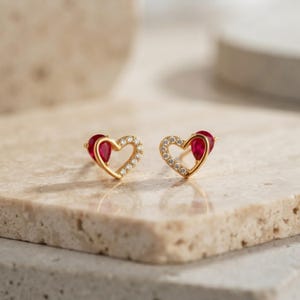 Puede incluir: Un par de pendientes de botón en forma de corazón de oro con una piedra roja en forma de lágrima en un lado del marco del corazón, acentuados con pequeñas piedras claras a lo largo del borde restante del contorno, creando una joya delicada.