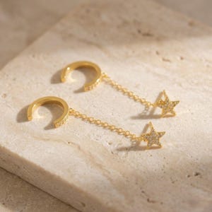 May include: A pair of gold-toned ear cuffs featuring a delicate chain connecting the cuff to a small, five-pointed star charm. Both the C-shaped ear cuff and the star charm are embellished with tiny, clear, sparkling stones set in the metal.