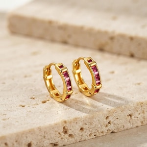 Peut inclure: Une paire de créoles dorées ornées d'une rangée de petites pierres précieuses rectangulaires rouge-rose foncé serties sur le bord extérieur de chaque anneau. Les boucles d'oreilles ont une finition métallique polie et un fermoir à charnière.