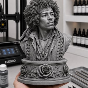 May include: A grey 3D printed bust of Jimi Hendrix featuring his signature afro hairstyle, a headband, and a military-style jacket. The figure is mounted on a circular pedestal base decorated with sculpted roses and intricate floral scrollwork.