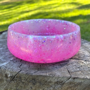 May include: A round, shallow bowl crafted from translucent pink resin. The material is embedded with a dense mixture of fine iridescent glitter and larger holographic flakes that catch the light, creating a sparkling, multi-dimensional effect.