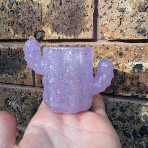 May include: A small, light purple cactus-shaped container made of a translucent resin material. The surface is embedded with numerous iridescent glitter particles that reflect light, creating a sparkling effect across the entire object.