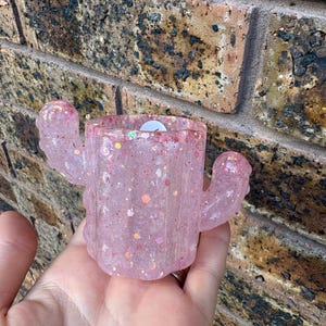 May include: A translucent pink cactus-shaped container crafted from resin, featuring embedded iridescent glitter and chunky sequins throughout its surface. The object has a cylindrical central body with two small, rounded arms extending from the sides.
