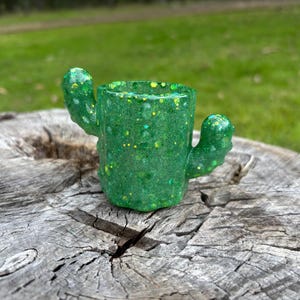 May include: A green cactus-shaped container made of resin with embedded yellow and green glitter throughout the body. The object features a cylindrical central section with two small, rounded arms extending from the sides, creating a distinct cactus silhouette.
