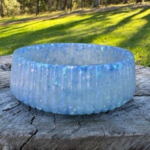 May include: A round, ribbed bowl with a translucent, light blue finish. The material is embedded with iridescent, shimmering flakes that catch the light, creating a multi-dimensional, sparkling effect across the entire exterior surface.