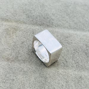 May include: A solid silver-toned ring featuring a thick, flat-topped square design. The band has a smooth, polished metallic surface with a reflective finish, showcasing a bold, geometric shape that wraps around the finger with a substantial profile.