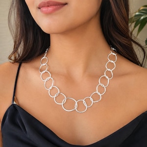 May include: A silver-toned necklace featuring a series of interconnected, irregular circular links. The chain is composed of varying sizes of open, rounded metal rings that form a continuous loop around the neck, creating a textured, link-style design.