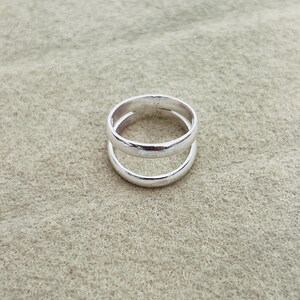 May include: A polished silver-coloured ring featuring a double-band design. The ring consists of two smooth, rounded bands connected at the sides, creating an open space between the upper and lower sections of the metal band structure.