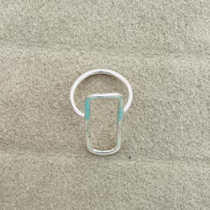 Puede incluir: Un anillo de tono plateado que presenta una banda circular conectada a un marco rectangular vertical. La superficie metálica tiene un acabado liso y pulido con sutiles acentos de color azul claro o turquesa visibles a lo largo de los bordes internos del rectángulo.
