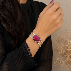 Peut inclure: Un bracelet manchette en argent orné d'une pierre précieuse magenta de forme ovale au centre. La pierre est sertie dans un cadre en argent perlé, et le bracelet se compose de deux fils d'argent parallèles qui se rejoignent au niveau du sertissage.