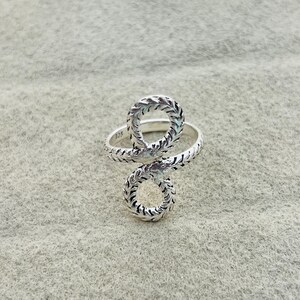 May include: A silver ring featuring a textured, rope-like design that forms a figure-eight or infinity shape. The metal surface has a polished finish with dark oxidised grooves that highlight the intricate, twisted pattern of the band.