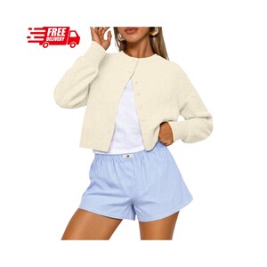 May include: A cream-colored long-sleeve knit cardigan with a round neckline and front button closure, worn over a plain white top and light blue and white pinstriped shorts. The cardigan features ribbed cuffs and a textured knit fabric construction.