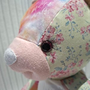 May include: A stuffed animal toy featuring a patchwork design with sections of pale yellow fabric printed with small pink flowers, sections of vibrant tie-dye fabric in pink, orange, and blue, and a solid light pink snout with a white nose patch.