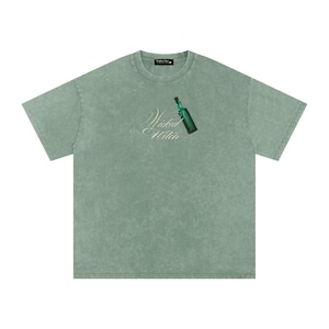 Puede incluir: Una camiseta de manga corta color verde salvia con acabado lavado mineral. El pecho presenta un gráfico de una pequeña botella de vidrio verde con tapón de corcho, acompañado del texto blanco en cursiva que dice Wicked the Witch.