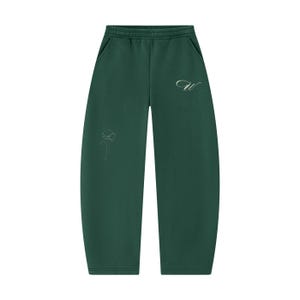 Puede incluir: Pantalones de chándal de color verde bosque oscuro con cintura elástica y bolsillos laterales. La parte delantera presenta una letra W cursiva plateada en el lado superior derecho y un delicado dibujo lineal plateado de una flor en la pierna izquierda.