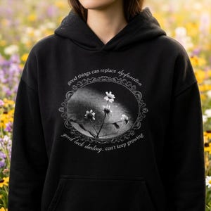 Pode incluir: Uma camisola preta com capuz apresentando um gráfico central de uma ilustração floral branca dentro de uma moldura oval ornamentada. O texto diz: good things can replace dysfunction, good luck darling, con't keep growing, em fonte branca.