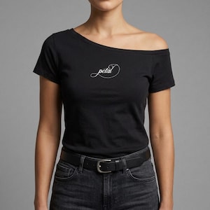 Puede incluir: Una camiseta negra de hombros descubiertos con la palabra petal escrita en una fuente cursiva blanca centrada en el pecho. La camiseta se combina con unos vaqueros de mezclilla gris oscuro sujetos por un cinturón de cuero negro con hebilla.