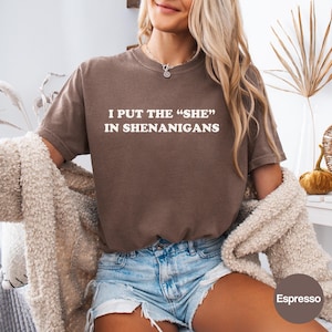 May include: A brown short-sleeve t-shirt featuring the white text I PUT THE "SHE" IN SHENANIGANS printed on the chest. The shirt has a crew neckline and a relaxed fit, paired with light blue denim shorts and a beige textured cardigan.