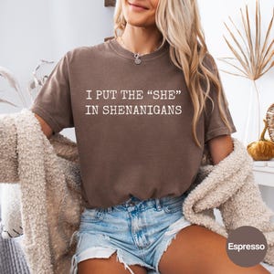 May include: A brown short-sleeved t-shirt featuring the white text I PUT THE "SHE" IN SHENANIGANS printed across the chest. The garment has a crew neckline and a relaxed fit, paired with light blue denim shorts and a textured beige cardigan.