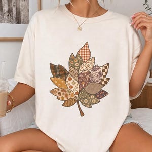 Pode incluir: Uma t-shirt de manga curta cor de creme com um grande gráfico central de uma folha de ácer. A folha está dividida em várias secções com padrões, incluindo xadrez, vichy, estampados florais, cogumelos, abóboras e uma pequena casa.