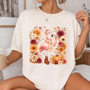 Puede incluir: Una camiseta de color crema con un gráfico de un flamenco rosa que lleva una bufanda, un lazo y botas marrones. El flamenco está rodeado por un arreglo cuadrado de flores en tonos otoñales naranja, amarillo y burdeos, junto a una calabaza.